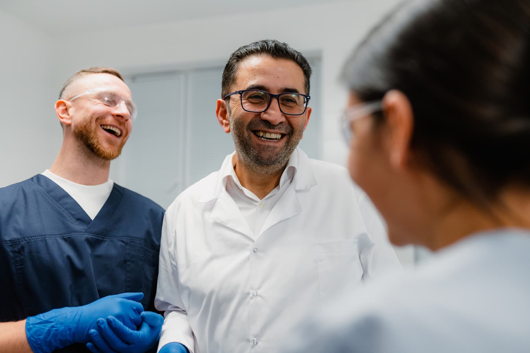 Group of healthcare professionals wearing lab coats, scrubs, and safety glasses laughing together in a clinical setting, highlighting teamwork, medical collaboration, and positive workplace culture.
