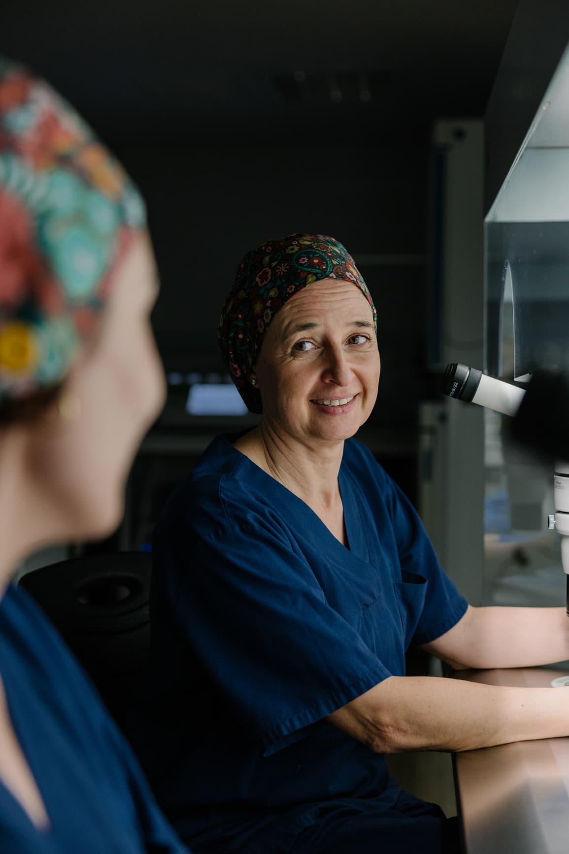 Smiling scientist wearing scrubs and a head covering while working at a microscope in a laboratory, representing medical research and scientific analysis.