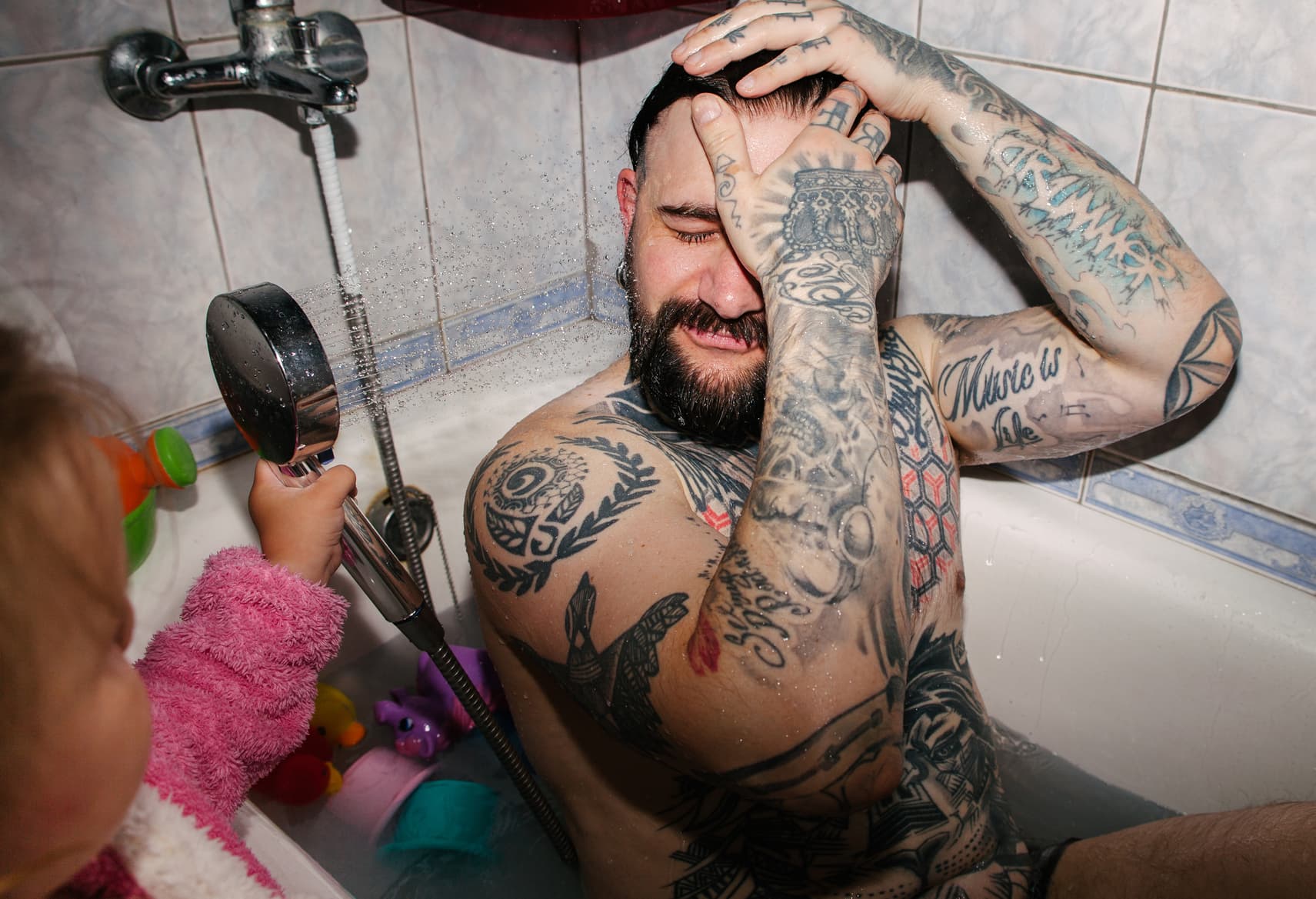 Child spraying water on a tattooed adult sitting in a bathtub, showing playful parenting, bath time interaction, and family humour.