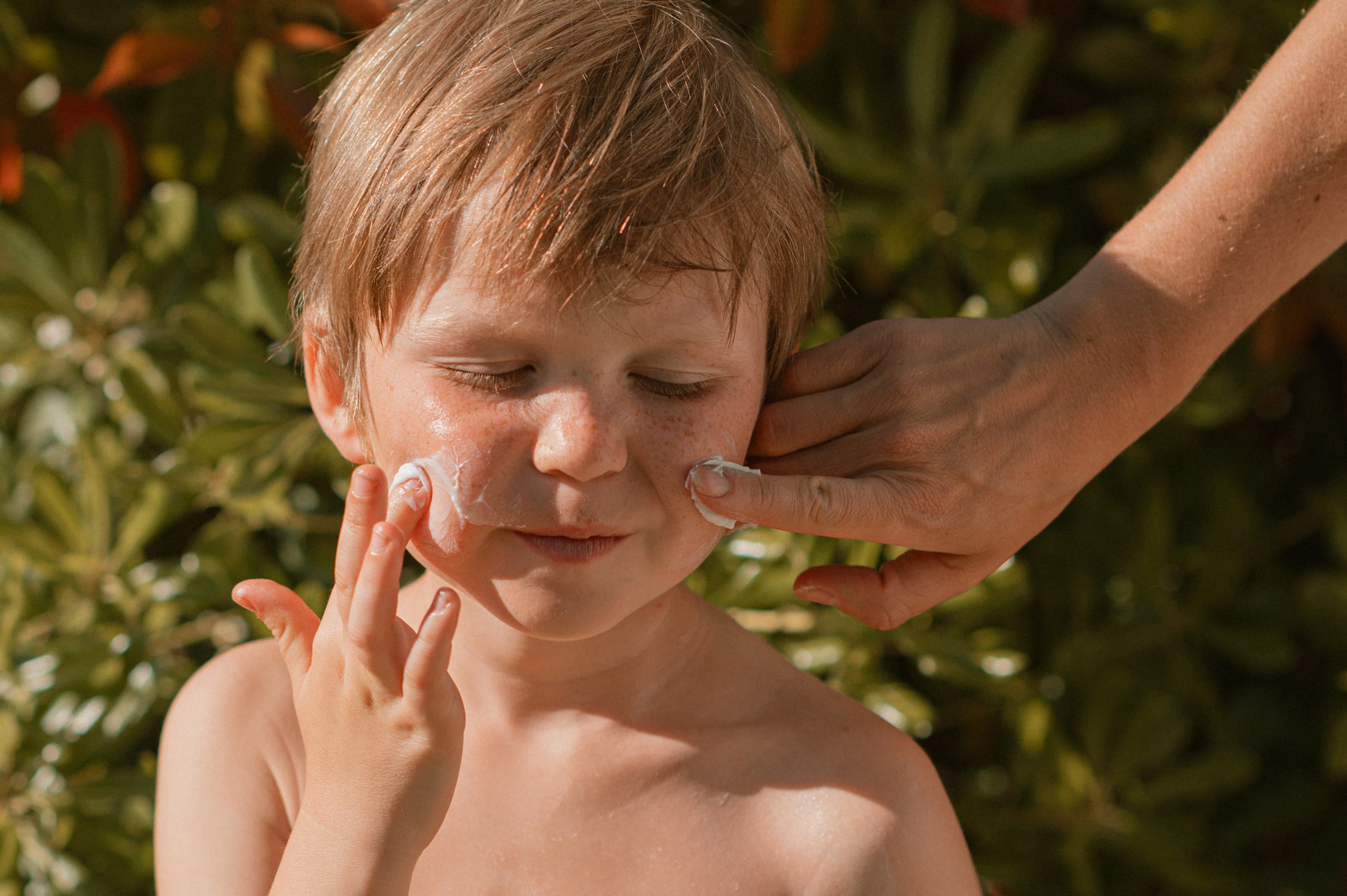 Child with freckles applying sunscreen to their face outdoors with adult assistance, emphasizing sun protection and child skincare in natural light.
