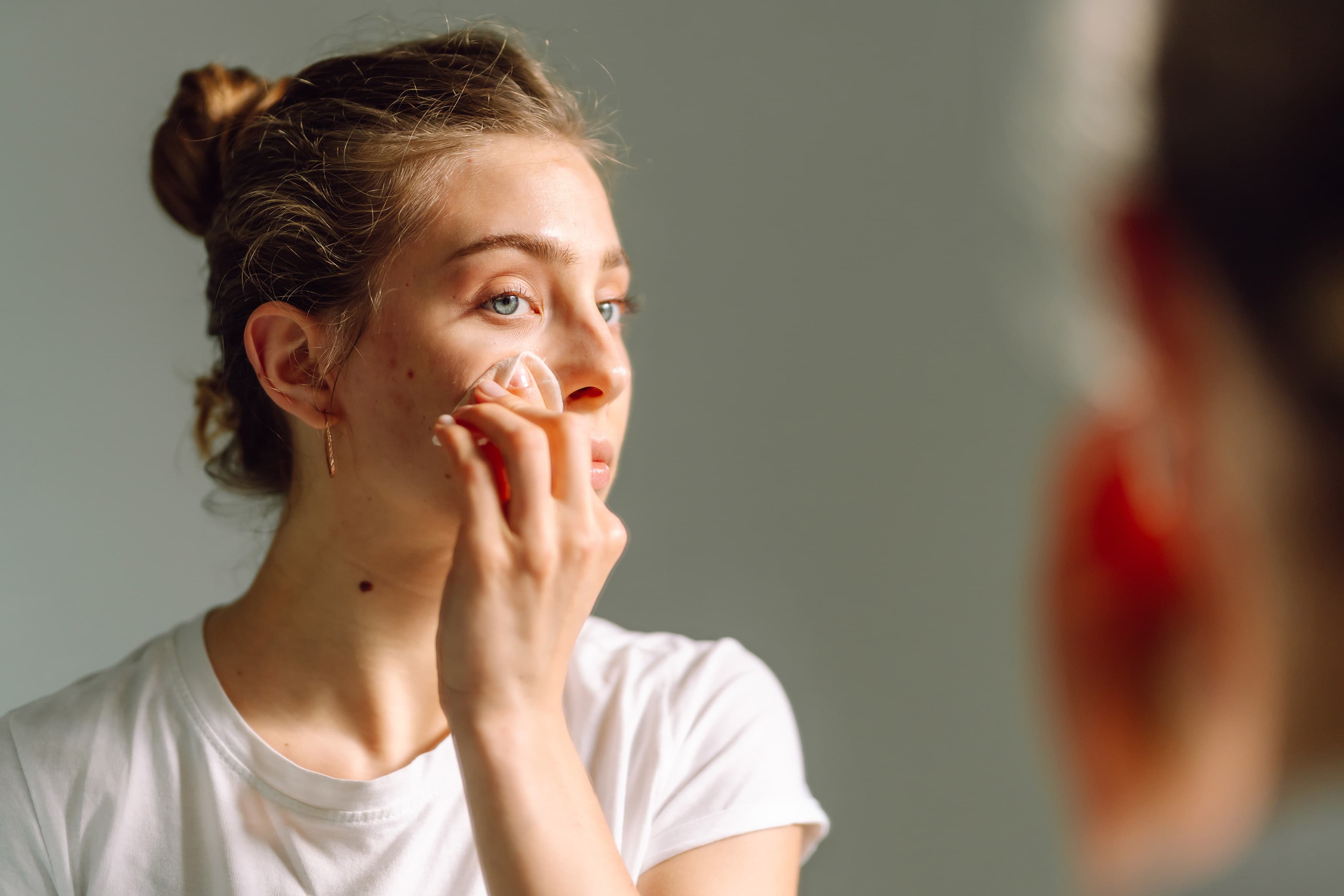 Young woman gently applying facial skincare product to her cheek while looking in a mirror, highlighting a daily skincare routine and natural skin texture.