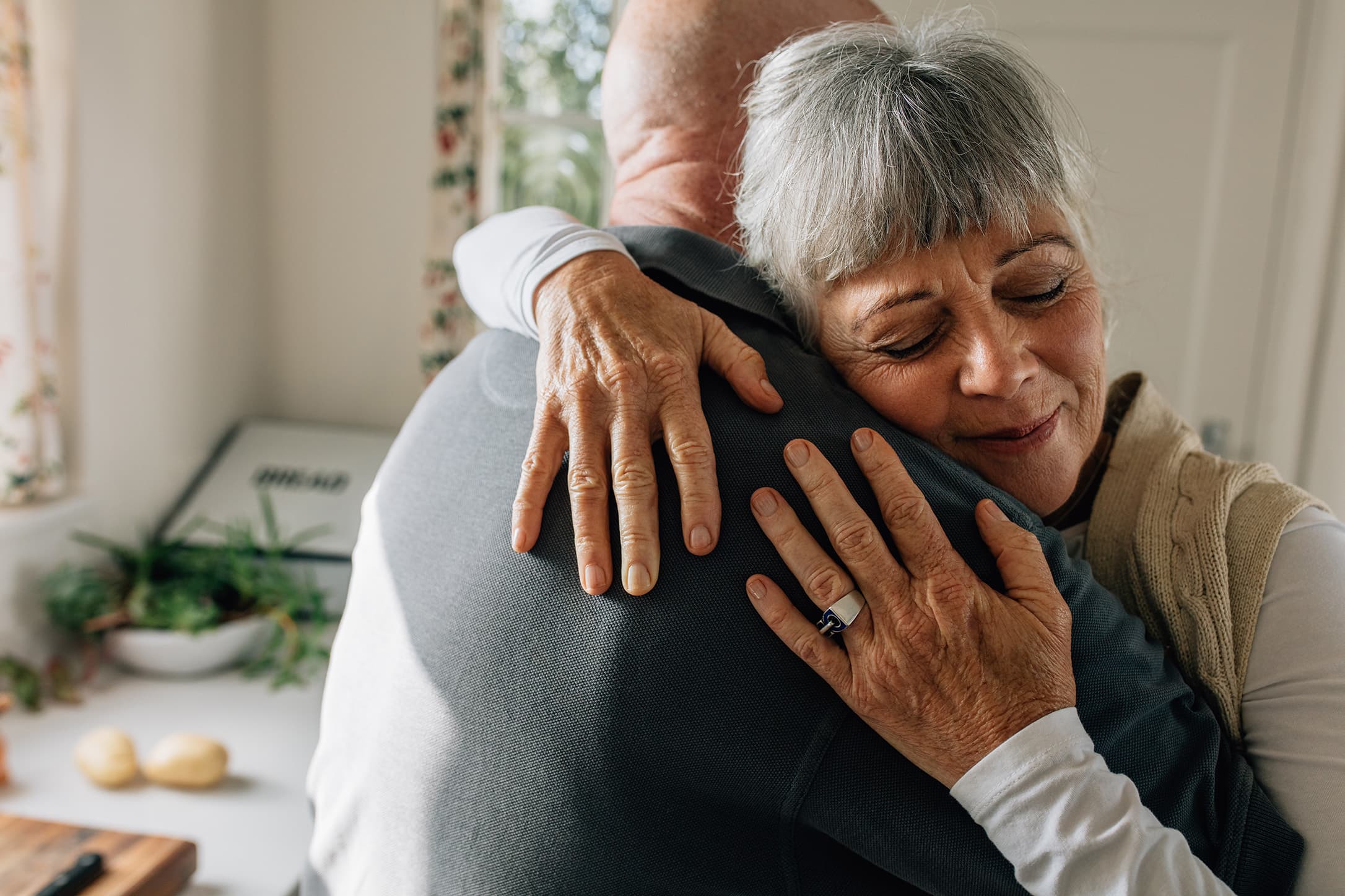 Older woman hugging her partner in a bright kitchen, expressing affection, emotional connection, and long-term companionship.