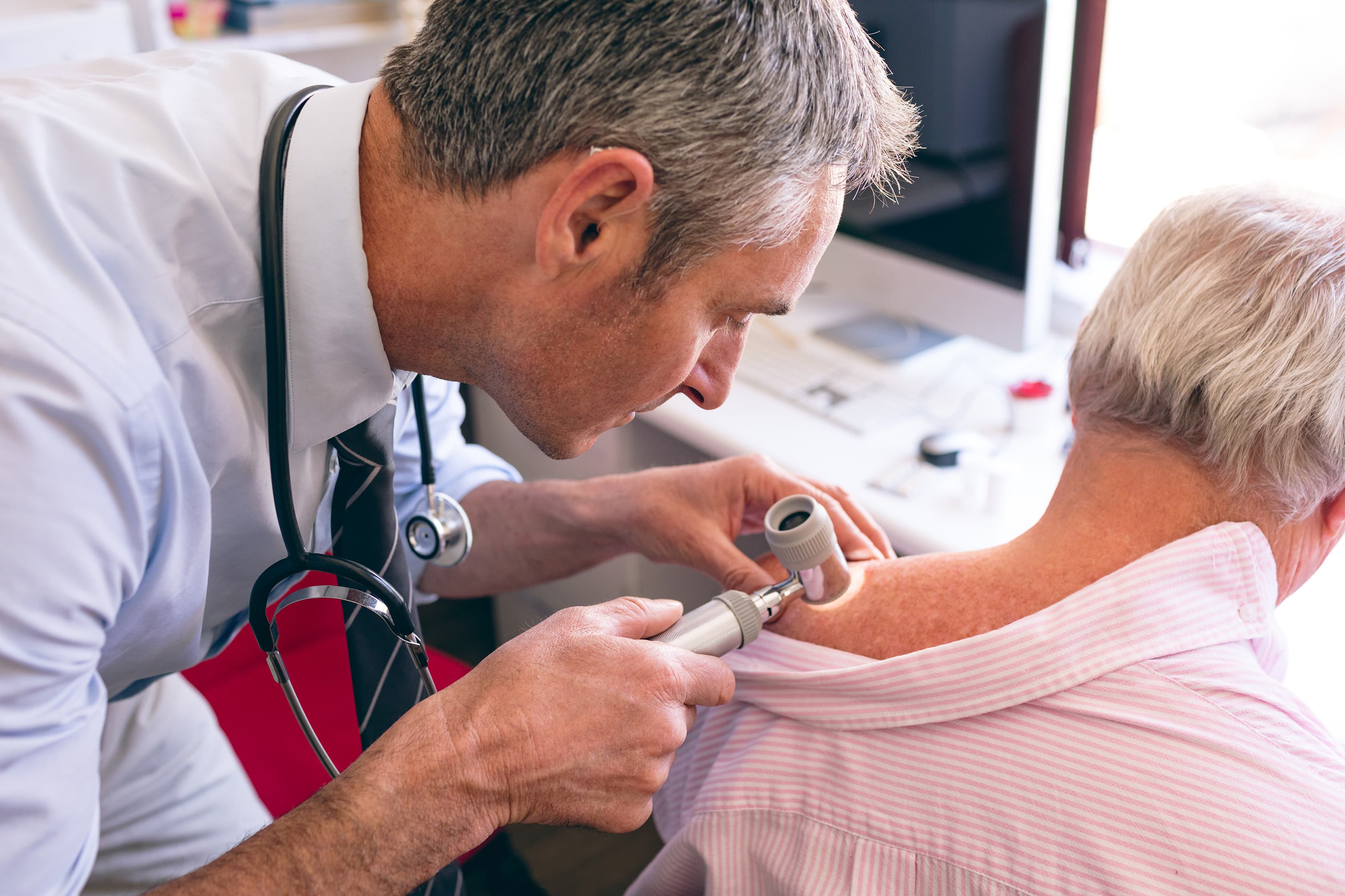 Doctor examining an older patient’s upper back with a dermatoscope during a medical skin check, representing preventive dermatology screening.