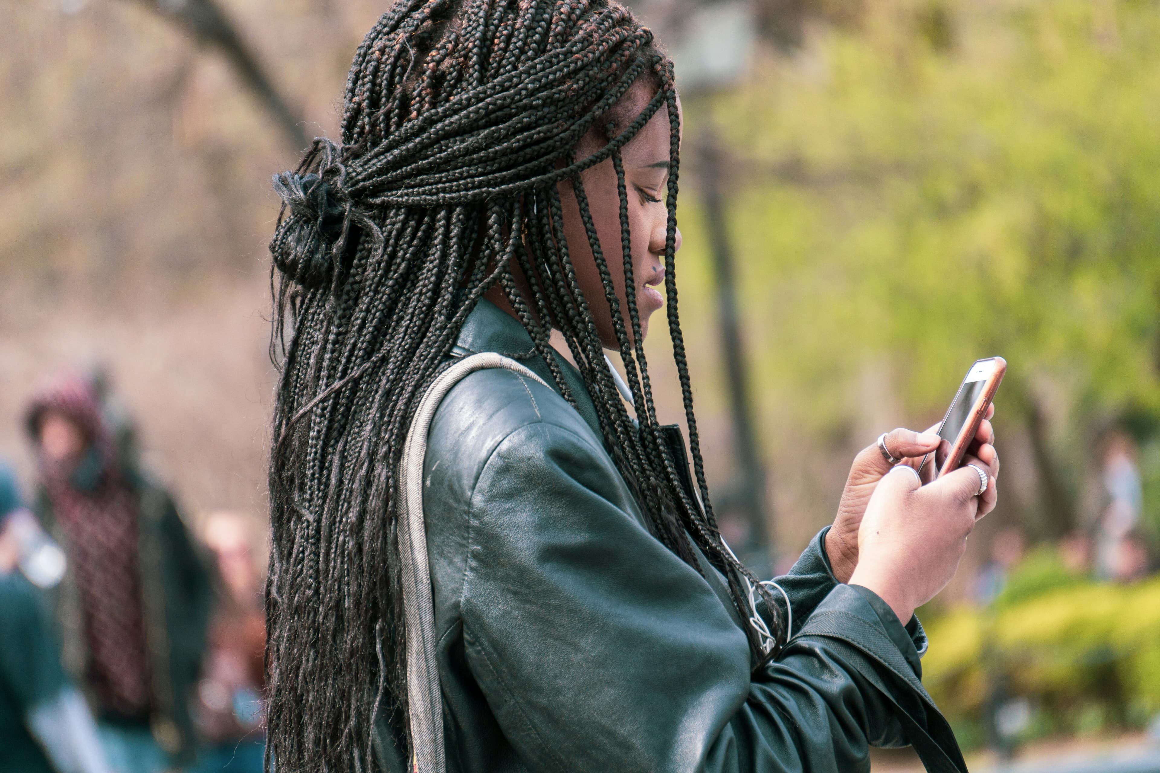Woman with long braided hair wearing a leather jacket while using a smartphone outdoors in a park, highlighting mobile technology use, everyday communication, and urban lifestyle.